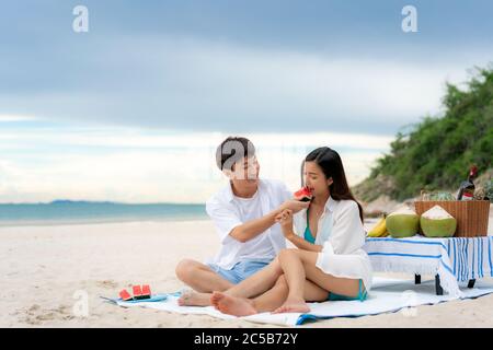 Jeune couple asiatique assis sur la couverture de pique-nique et mangeant de la pastèque sur la plage et près de la mer avec des fruits tropicaux en arrière-plan. Été, vacances, Banque D'Images