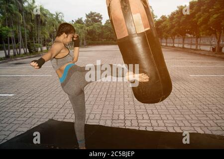 Belle jeune fille blanche asiatique avec de longs cheveux de 20-30 ans. Pratiquez la boxe thaï de Muay en donnant un coup de pied au sac de sable de boxe dans le parc. Banque D'Images