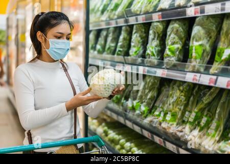une jeune femme asiatique choisit des légumes tout en faisant ses courses de nourriture dans un supermarché et porte un masque médical pour prévenir la pandémie du coronavirus (covid-19). n Banque D'Images