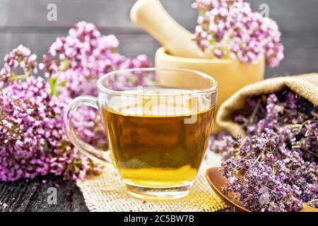 Thé à base de plantes origan dans une tasse de verre sur une serviette en toile de jute, fleurs fraîches dans le mortier et sur la table, fleurs de marjolaine séchées dans un sac et cuillère sur un sanglier en bois Banque D'Images