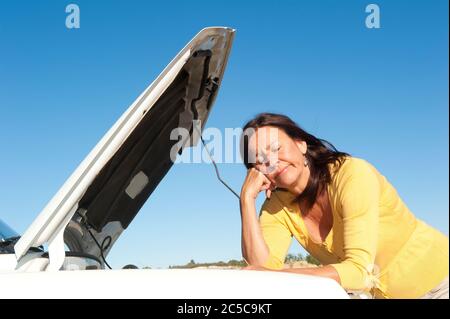 Femme mûre stressée en panne avec voiture sur route éloignée en attente d'assistance, pour l'aide, isolé avec ciel bleu comme arrière-plan et espace de copie. Banque D'Images