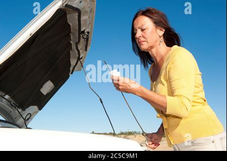 Femme mûre stressée en panne avec voiture sur route éloignée, en vérifiant l'huile et en attente d'assistance, pour l'aide, isolée avec ciel bleu comme fond et co Banque D'Images