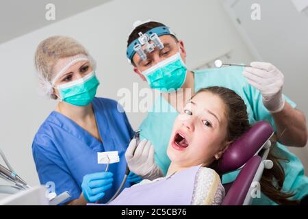 Portrait de peur teen girl avec la bouche ouverte pendant un traitement dentaire avec deux dentistes debout près de présidence Banque D'Images