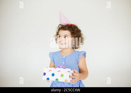 Jolie petite fille souriante avec cheveux fclés foncés dans la robe bleue et le chapeau d'anniversaire regardant avec joie de côté tout en tenant la boîte cadeau sur fond blanc Banque D'Images