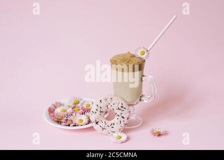 Café de Dalgona dans une tasse transparente avec un tube en papier, une assiette avec des fleurs pâquerettes et un délicieux donat arrosoir avec glaçage rose Banque D'Images