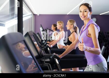 Smiling athletic girls running sur tapis roulant dans fitness club giving Thumbs up Banque D'Images