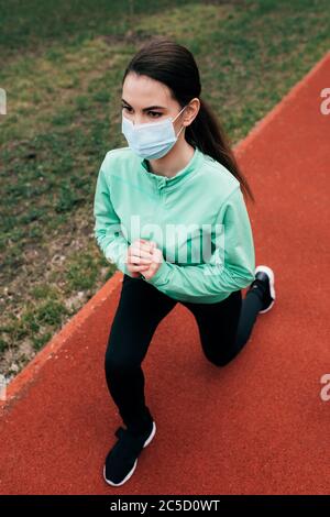 Vue en grand angle de sportswoman dans un masque médical faisant des fentes sur la piste de course pendant l'exercice en plein air Banque D'Images