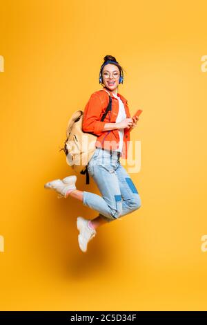 élève enthousiaste dans un casque et des lunettes sautant tout en tenant le smartphone sur fond jaune Banque D'Images