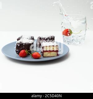 Gâteaux traditionnels de lamington australiens avec confiture de fraise, chocolat et noix de coco. Contre d'un verre d'eau, une éclaboussure d'eau, chute Banque D'Images