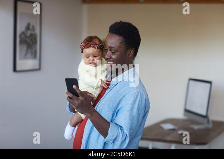 Père souriant africain tenant la petite fille sur ses mains et utilisant son téléphone portable à la maison Banque D'Images