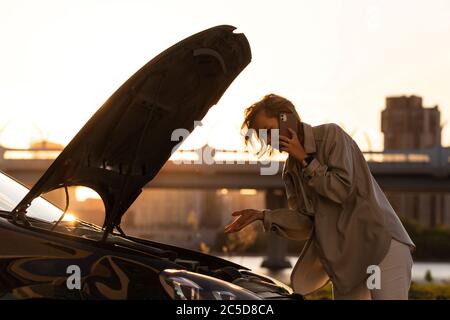 Une femme sans défense, chauffeur appelant pour obtenir de l'aide ou de l'aide, qui a regardé une voiture en panne, s'est arrêtée au bord de la route. Ne comprend pas ce qui s'est passé. Heure du coucher du soleil. Banque D'Images