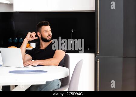beau indépendant regardant loin tout en étant assis près d'un ordinateur portable Banque D'Images