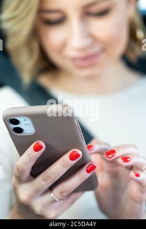 Mise au point logicielle sélective sur téléphone mobile, gros plan. Femme souriante floue assise en voiture sur le siège du passager fixé par une ceinture de sécurité, à l'aide d'un smartphone, Banque D'Images