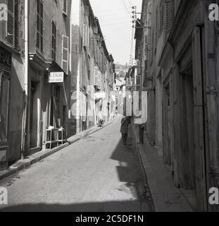 Années 1950, été et dans une rue étroite, un commerçant français dans son tablier et sa brosse parlant avec un homme du coin, Nice, Sud de la France. Banque D'Images