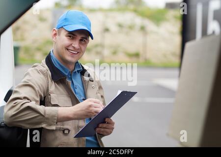 Portrait d'un jeune liveur remplissant des documents et souriant à l'appareil photo tout en se tenant près de la voiture à l'extérieur Banque D'Images