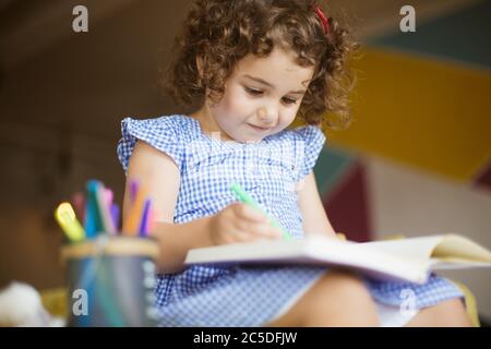 Belle petite fille avec cheveux maurités foncés dans la robe dessin rêveur dans le livre de coloriage avec stylos-feutre sur le canapé à la maison Banque D'Images