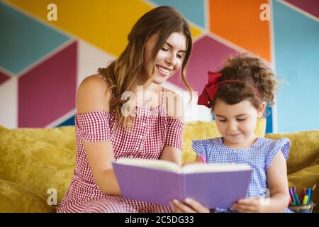 Belle mère souriante avec sa petite fille gentiment dessiner dans un livre avec des stylos-feutres tout en passant du temps ensemble sur le canapé à la maison Banque D'Images