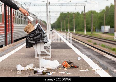 La main d'un inconnu jette des déchets dans un panier de déchets surpeuplé sur une plate-forme de chemin de fer. Poubelle. Une pile de déchets en plastique sur le sol. Banque D'Images