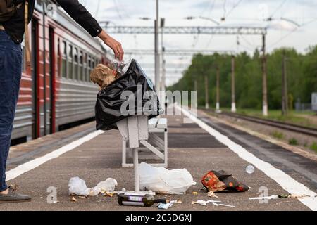 La main d'un inconnu jette des déchets dans un panier de déchets surpeuplé sur une plate-forme de chemin de fer. Poubelle. Une pile de déchets en plastique sur le sol. Banque D'Images