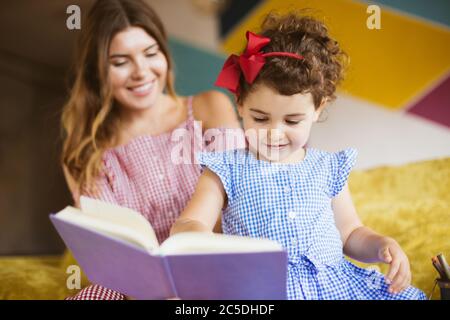 Jolie petite fille avec des cheveux de curly foncé dans la robe lire heureusement livre d'enfant avec la mère sur le canapé à la maison Banque D'Images