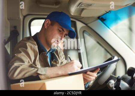 Jeune messager remplissant le formulaire en étant assis dans la voiture qu'il livrant le paquet Banque D'Images