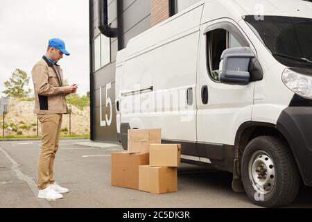 Jeune homme en uniforme utilisant son téléphone portable tout en se tenant près de la camionnette et des colis à l'extérieur près de l'entrepôt Banque D'Images