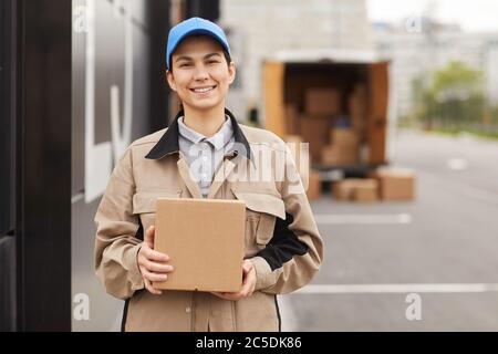 Portrait de la jeune femme en uniforme tenant le colis et souriant à la caméra tout en se tenant près de l'entrepôt Banque D'Images
