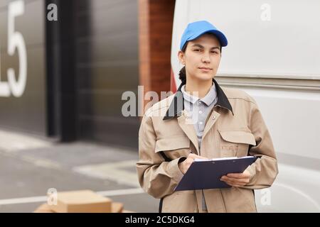 Portrait de la jeune femme en uniforme remplissant la forme en se tenant à l'extérieur, elle travaille dans l'entrepôt de livraison Banque D'Images