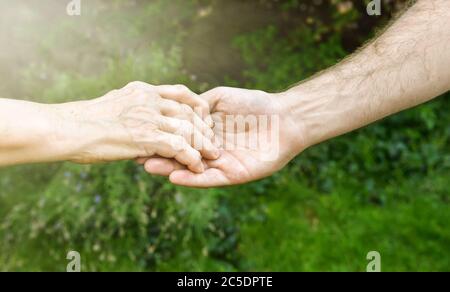 Les mains des aînés et des jeunes à l'extérieur. Femme âgée et jeune homme tenant les mains ensemble, fond vert flou, lumière du soleil. Amour, chaleur, prendre la voiture Banque D'Images