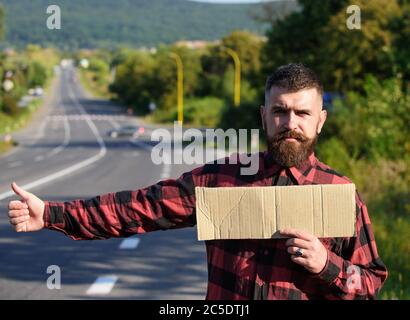 Homme au visage et à la barbe stricts voyageant en randonnée avec la route sur le fond. Concept de voyage et de randonnée. Taille-culotte essayez d'arrêter la voiture avec une affiche en carton et un geste de pouce vers le haut, espace de copie. Banque D'Images