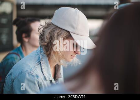 Belle fille blonde dans un chapeau avec les cheveux courts est assis dans un groupe de filles, regarde vers le bas. Solitude entre amis. Mise au point sélective. Banque D'Images