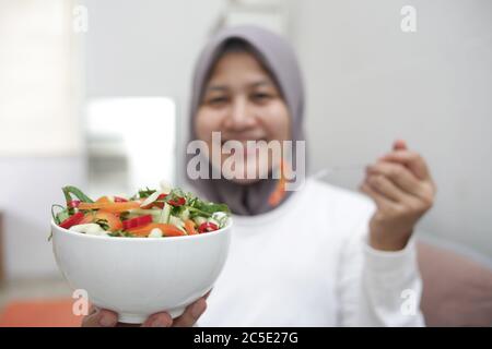 Femme asiatique musulmane portant hijab manger une nourriture saine, salade de légumes verts, après l'exercice à la maison, garder en bonne santé et en forme pendant le nouveau mode de vie normal co Banque D'Images