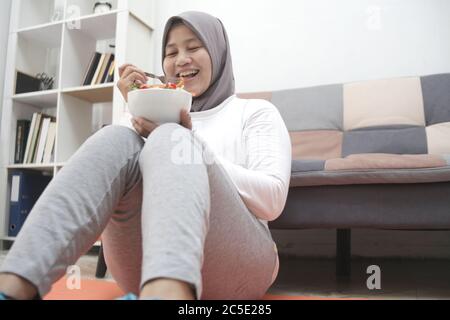 Femme asiatique musulmane portant hijab manger une nourriture saine, salade de légumes verts, après l'exercice à la maison, garder en bonne santé et en forme pendant le nouveau mode de vie normal co Banque D'Images