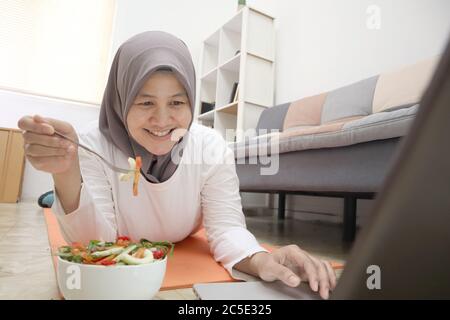 Femme asiatique musulmane portant hijab manger une nourriture saine, salade de légumes verts, après l'exercice à la maison, garder en bonne santé et en forme pendant le nouveau mode de vie normal co Banque D'Images