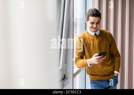 Jeune homme joyeux tenant un téléphone portable et regardant l'appareil photo sur le fond du bureau Banque D'Images