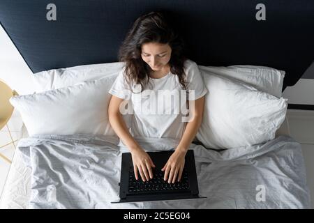 Belle fille brunette au lit avec un ordinateur portable. Travail du matin à tard en soirée. Gadgets électroniques avant le coucher ou le matin Banque D'Images