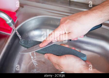 Gros plan des mains d'un homme affûtez soigneusement manuellement un couteau sous un jet d'eau sur une pierre à affûter. Aiguisage domestique. Entretien des couverts de cuisine. Banque D'Images