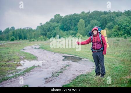 Un randonneur touristique se promène le long de la route de terre avec un sac à dos dans une veste rouge sous la pluie Banque D'Images