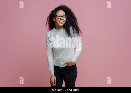 Heureuse et joyeuse femme afro-américaine a une expression insouciante heureuse, tient la main dans la poche et regarde de côté avec un large sourire, porte blanc turtleneck A. Banque D'Images