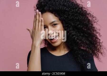 Bonne adolescente a du plaisir et cache le visage avec la paume, a l'expression joyeuse, les cheveux bouclés, vêtus de vêtements noirs décontractés, isolé sur le dos rose Banque D'Images