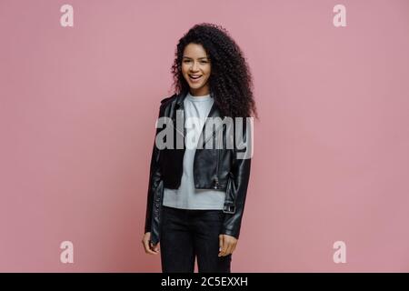 Bonne positive Afro-américaine femme vêtue de cuir veste, prête pour la marche en plein air, pose sur fond rose. Joyeux GI millénaire à la peau sombre Banque D'Images