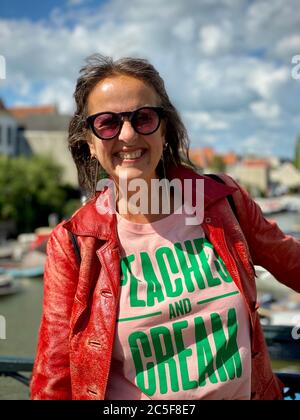 Portrait d'une femme mature et plus jeune, portant des lunettes de soleil et une veste en cuir rouge vintage, adossée à une main courante, montrant l'émotion souriante. Banque D'Images