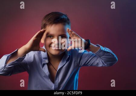 Le portrait studio d'un garçon souriant est un brunette dans une chemise bleue sur fond violet et marron. Tenant ses mains sur son visage Banque D'Images