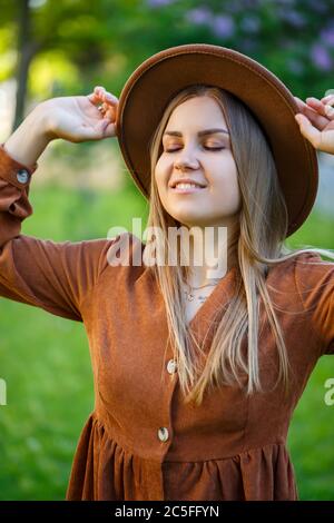 Une belle fille aux cheveux blonds tient un chapeau dans ses mains et se tient avec ses yeux fermés près d'un buisson lilas. Jeune femme dans un jardin avec un être en fleurs Banque D'Images