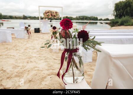 Fleurs de dahlia rouge foncé dans un vase en verre transparent. Cérémonie de mariage en plein air sur une plage de sable. Concept de mariage. Banque D'Images