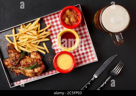 Ailes de poulet cuites avec frites sur un plateau en fer avec verre de bière sur fond noir Banque D'Images