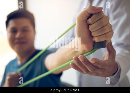 Gros plan de la main patient faisant de l'exercice avec un bracelet d'exercice flexible et une main de thérapeute physique pour aider dans la salle de clinique. Banque D'Images