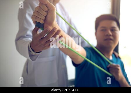 Gros plan de la main patient faisant de l'exercice avec un bracelet d'exercice flexible et une main de thérapeute physique pour aider dans la salle de clinique. Banque D'Images