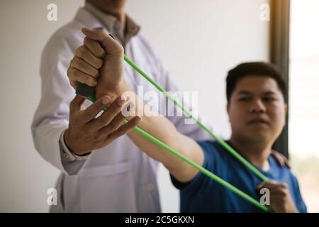 Gros plan de la main patient faisant de l'exercice avec un bracelet d'exercice flexible et une main de thérapeute physique pour aider dans la salle de clinique. Banque D'Images