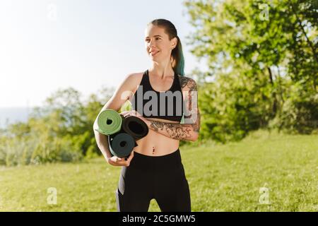 Portrait de femme souriante en haut noir sportif et leggings debout avec des tapis de yoga dans les mains et regard rêveur de côté avec belle vue sur Banque D'Images